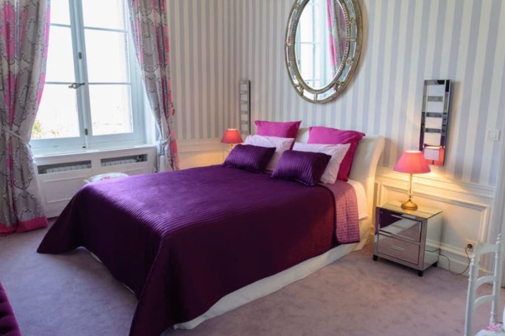 A cozy bedroom with striped wallpaper, purple bedding, and elegant furnishings offers a serene retreat for travelers seeking comfort and style in their accommodations.