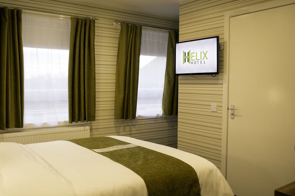 A cozy hotel room features a neatly made bed with green accents, striped walls, and a mounted TV displaying "Helix Hotel," ideal for travelers seeking comfort and modern amenities.