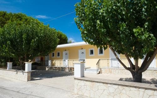 A serene Mediterranean villa with yellow walls, white shutters, and lush greenery offers a tranquil retreat, ideal for travelers seeking relaxation in a picturesque coastal setting.