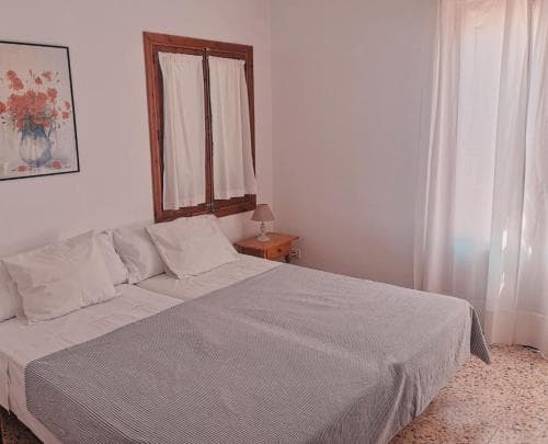 A cozy bedroom with a neatly made bed, wooden-framed window, soft curtains, and floral artwork creates a serene atmosphere ideal for travelers seeking a peaceful retreat.