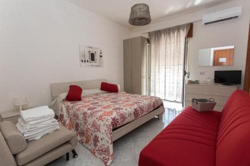 A cozy hotel room features a neatly made bed with a floral-patterned duvet, a red sofa, and a balcony door letting in soft light, ideal for travelers seeking comfort and relaxation.