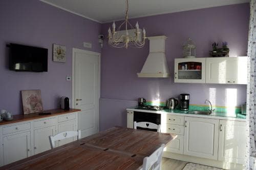 A cozy kitchen with lavender walls, white cabinetry, and a rustic wooden dining table creates an inviting atmosphere, perfect for travelers seeking a charming and homey accommodation experience.