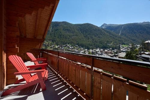 A cozy wooden balcony with red chairs overlooks a picturesque mountain village under a clear blue sky, ideal for travelers seeking relaxation in scenic alpine settings.