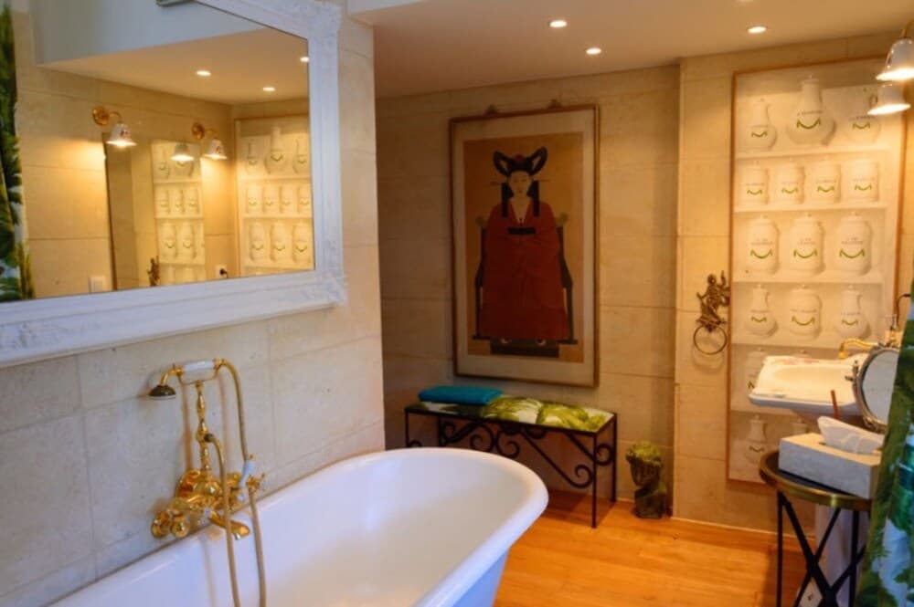 A luxurious bathroom features a clawfoot tub with gold fixtures, a large mirror, an artistic portrait on the wall, and shelves displaying decorative jars, creating an elegant and serene atmosphere ideal for high-end accommodation marketing.