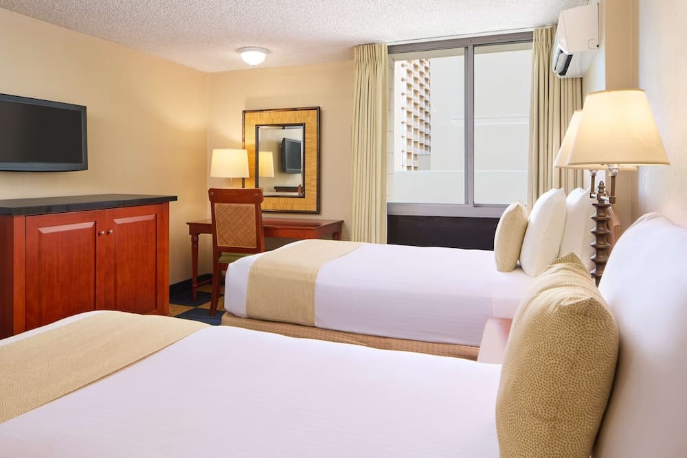 A cozy hotel room features two neatly made beds, a wooden desk with a chair, a mounted TV, and a large window with city views, ideal for travelers seeking comfort and convenience.