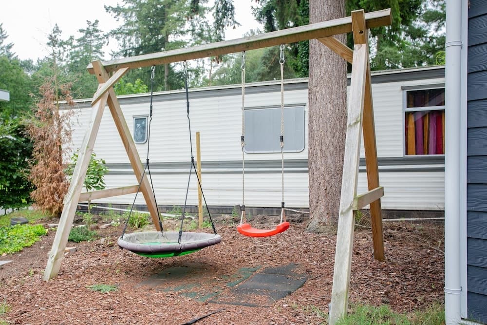 A cozy backyard swing set with colorful seats stands invitingly near a mobile home amidst lush greenery, perfect for family vacations or weekend getaways.