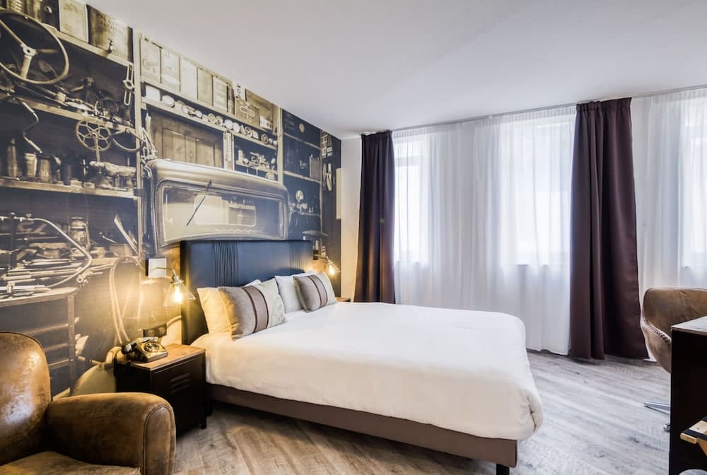 A cozy hotel room features a large bed with plush pillows, a vintage-themed wall mural, leather armchairs, and soft lighting, creating an inviting atmosphere ideal for travelers seeking comfort and style.