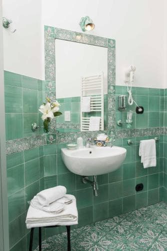 A serene hotel bathroom features vibrant green tiles, a white sink, neatly folded towels, and a decorative mirror, ideal for travelers seeking a refreshing and stylish accommodation experience.