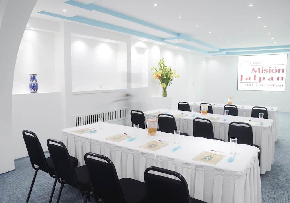 A serene conference room with white tables, black chairs, floral arrangements, and a presentation screen displaying "Misión Jalpan," ideal for professional meetings or workshops.