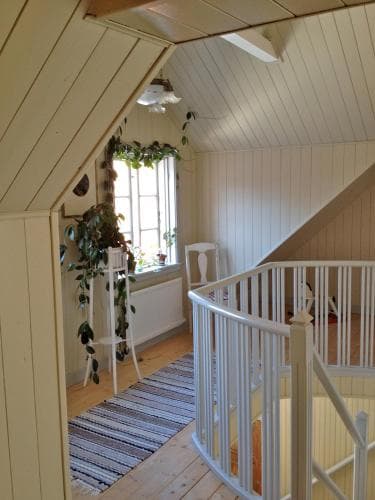 A cozy attic space with white wooden walls, a spiral staircase, and a window adorned with greenery creates a charming and inviting atmosphere, perfect for travelers seeking a quaint and serene accommodation.