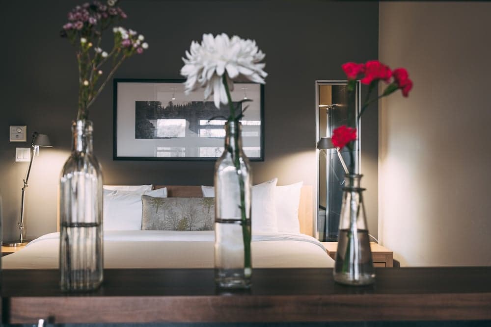 A cozy hotel room exudes tranquility with its neatly made bed, soft lighting, and vibrant flowers in glass vases on the dresser, ideal for travelers seeking relaxation and comfort.