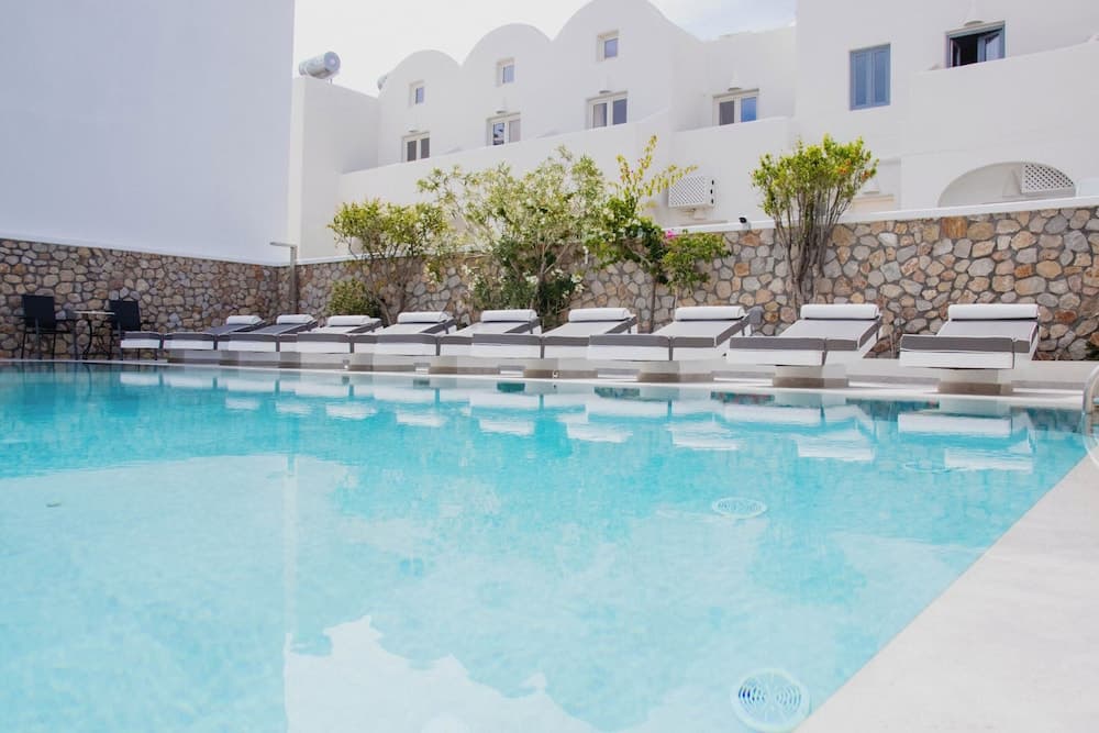 A serene resort pool area features crystal-clear turquoise water surrounded by white lounge chairs, lush greenery, and stone walls, ideal for travelers seeking relaxation in a Mediterranean-style setting.