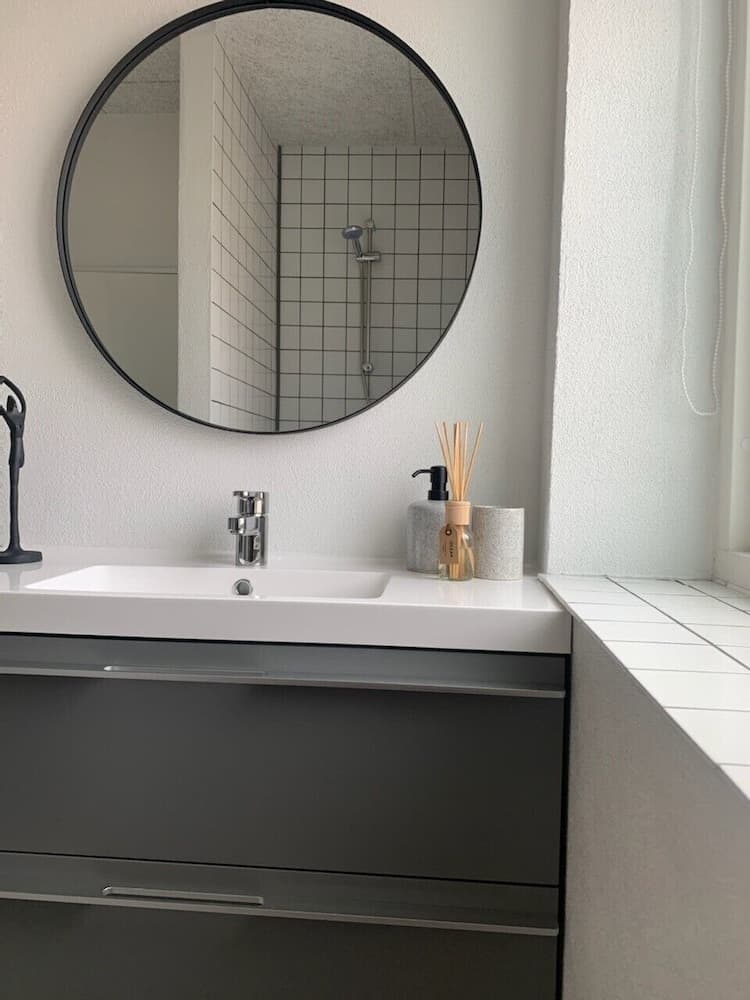 A modern bathroom features a sleek black vanity, white countertop, and a round mirror reflecting a tiled shower area, creating a clean and minimalist atmosphere ideal for travelers seeking stylish accommodations.