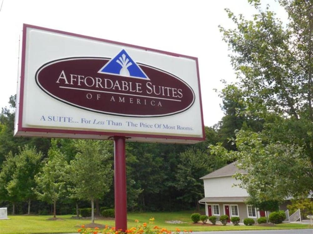 A welcoming roadside sign for Affordable Suites of America stands amidst lush greenery, inviting travelers to enjoy budget-friendly accommodations in a serene suburban setting.