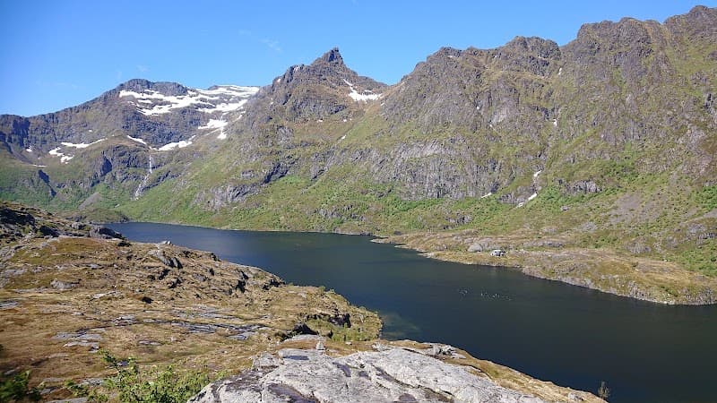 A serene mountain lake surrounded by rugged peaks under a clear blue sky offers an ideal backdrop for adventure seekers exploring the natural beauty of Moskenes, Norway.