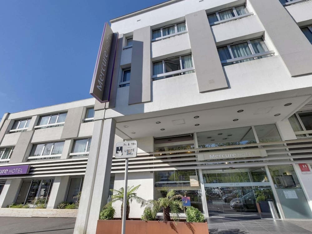 A modern Mercure Hotel entrance features sleek white architecture, glass doors, and potted plants under a clear blue sky, ideal for travelers seeking comfortable accommodations in an urban setting.