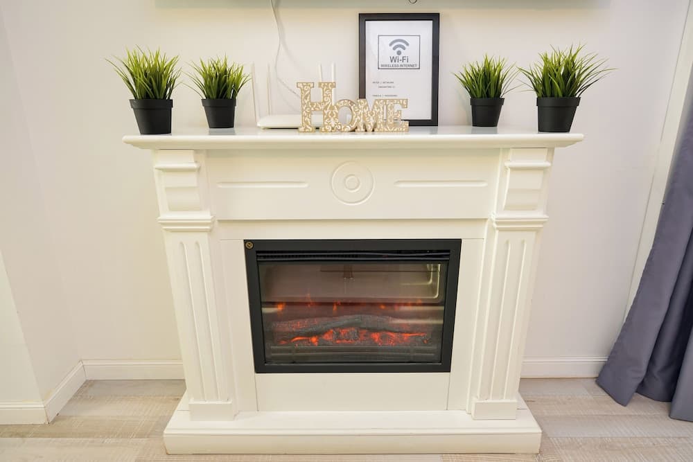 A cozy indoor setting features a white electric fireplace adorned with potted plants, decorative "HOME" letters, and a Wi-Fi sign, ideal for travelers seeking warmth and comfort in a modern living space.