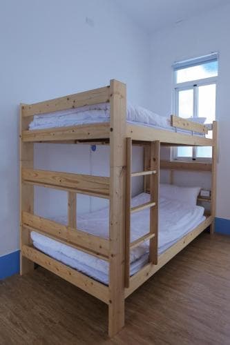 A cozy hostel room features a wooden bunk bed with white linens under soft natural light from a window, ideal for budget travelers seeking simple accommodations.