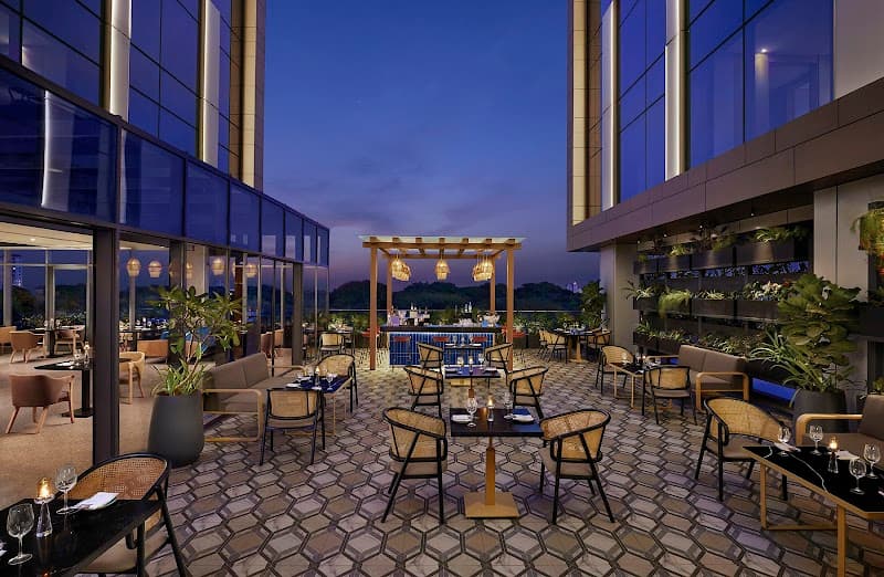 A serene rooftop dining area at Hyatt Centric Hebbal Bengaluru features modern tables, cozy chairs, lush greenery, and a stunning cityscape backdrop, ideal for travelers seeking a luxurious evening ambiance.