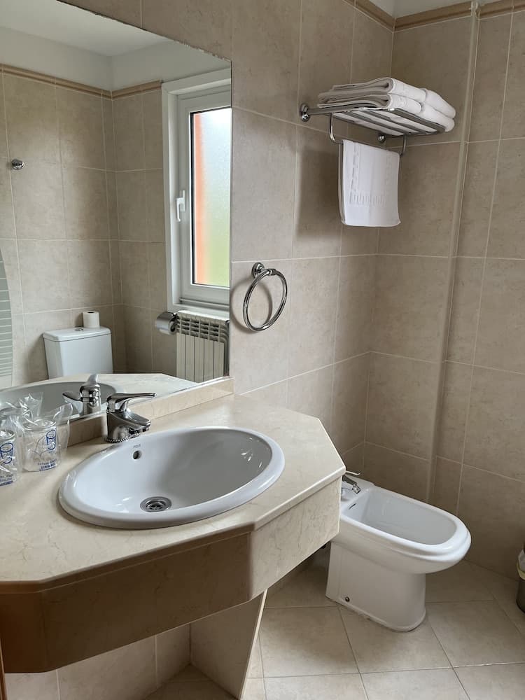 A cozy hotel bathroom features beige tiled walls, a sleek sink vanity, a bidet, neatly folded towels on a rack, and a frosted window, ideal for travelers seeking comfort and cleanliness.