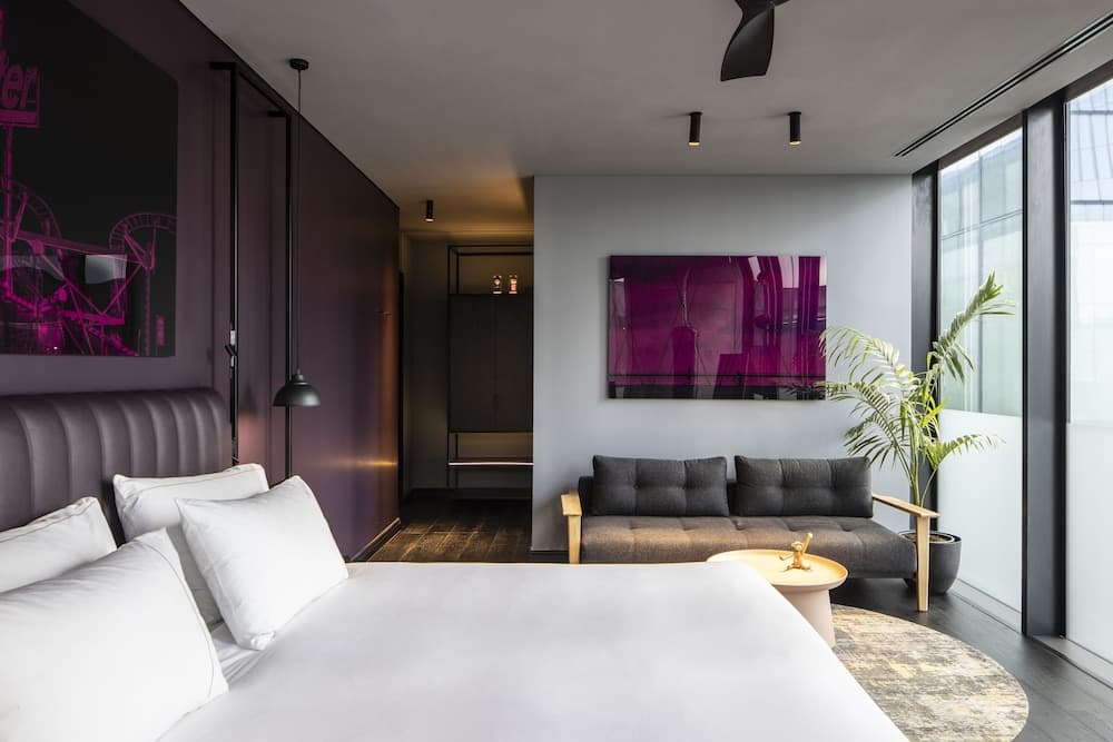 A modern hotel room features a cozy bed, sleek furnishings, vibrant purple artwork, and large windows, creating an inviting atmosphere ideal for travelers seeking comfort and style.