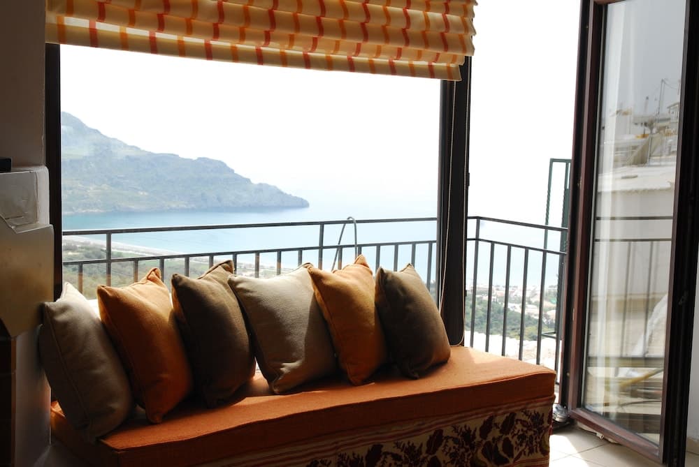 A cozy balcony bench adorned with earth-toned cushions overlooks a serene coastal landscape, perfect for travelers seeking relaxation and scenic views.