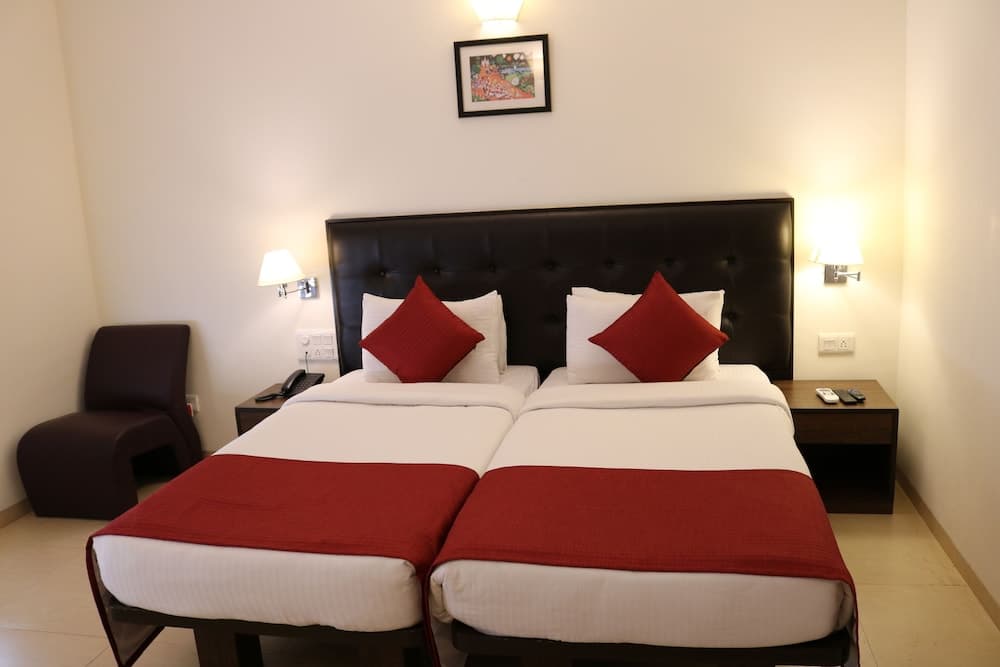 A cozy hotel room features two neatly arranged twin beds with red accents, a plush armchair, and warm wall-mounted lamps, ideal for travelers seeking comfort and relaxation.
