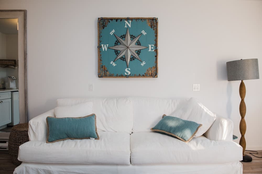 A cozy living room features a white sofa adorned with teal pillows under a rustic compass wall art, creating a serene nautical vibe ideal for vacation rental interiors.
