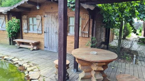 A cozy wooden cabin with rustic shutters and a charming outdoor seating area by a tranquil pond invites travelers seeking a serene retreat in nature.