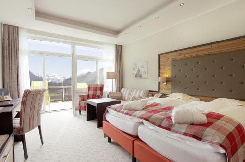 A cozy hotel room with a scenic mountain view through large windows, featuring a plush bed with red plaid bedding, modern furnishings, and a warm, inviting atmosphere ideal for travelers seeking relaxation in a picturesque alpine setting.