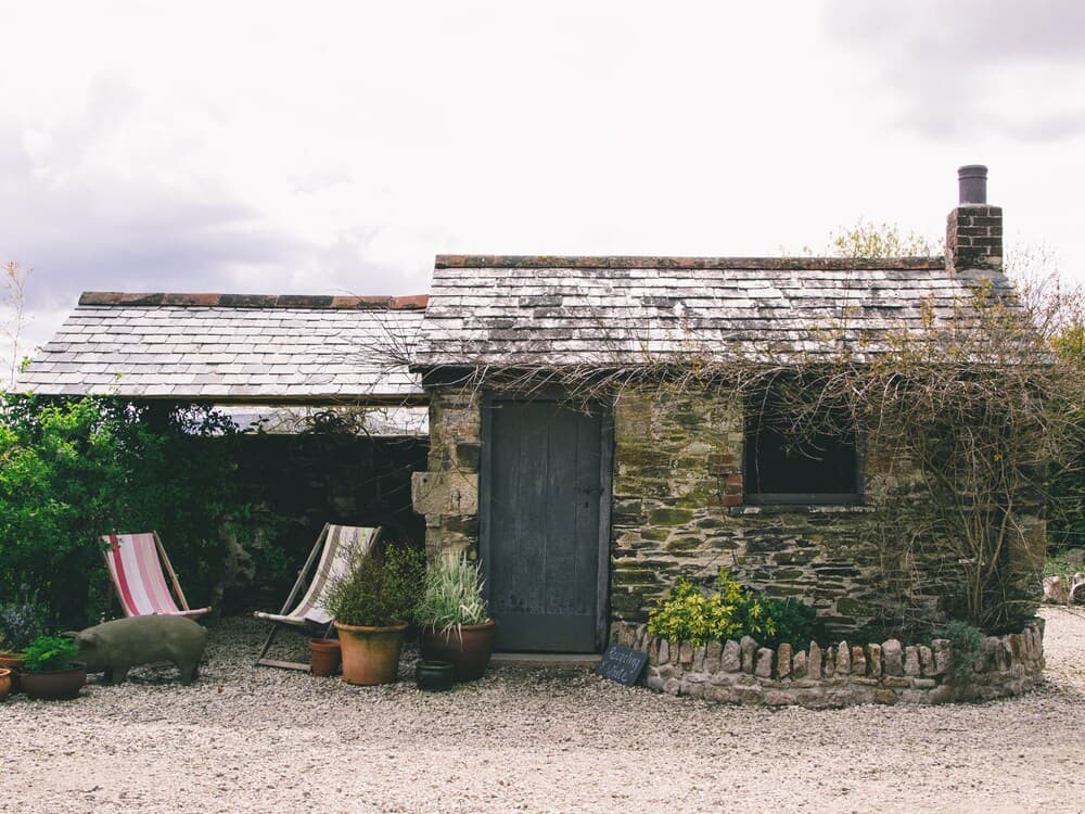 A quaint stone cottage with a rustic charm, surrounded by potted plants and garden furniture, offers a serene retreat for travelers seeking a peaceful countryside escape.