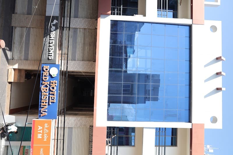 A vibrant urban scene featuring the Hotel Lord Krishna in Dwarka, Gujarat, with its modern facade, reflective glass panels, and nearby ATM signage, ideal for travelers seeking accommodation and convenience in a bustling city environment.