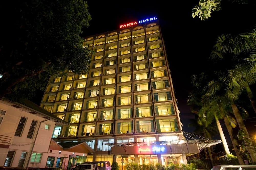 A warmly lit hotel building stands tall at night, surrounded by lush tropical foliage, creating an inviting atmosphere ideal for travelers seeking accommodation in a serene urban setting.