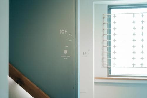 A serene indoor hallway features a teal wall with floor indicators and a window adorned with cross-patterned blinds, ideal for travelers seeking modern architectural details in urban accommodations.