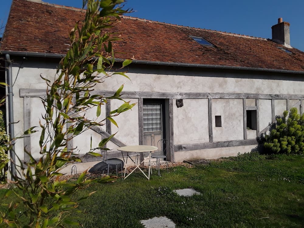 A quaint rural cottage with a rustic red-tiled roof and wooden-framed windows sits amidst lush greenery, featuring a cozy outdoor seating area perfect for a peaceful getaway.