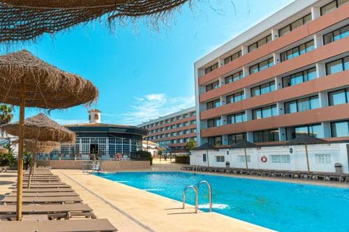 A vibrant resort scene features a sparkling pool lined with thatched umbrellas and lounge chairs under a clear blue sky, set against the backdrop of a modern hotel building, ideal for travelers seeking relaxation and leisure.