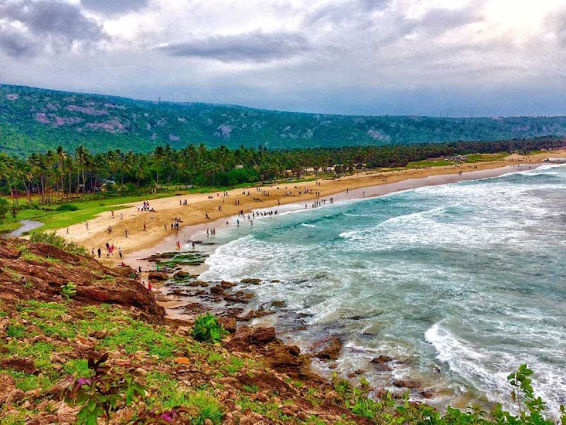 A serene beach scene at Yarada Beach in Andhra Pradesh, India, features turquoise waters lapping against sandy shores lined with palm trees, under a moody sky, ideal for travelers seeking relaxation and natural beauty.