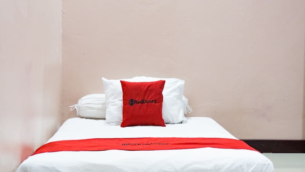 A cozy budget hotel room features a neatly made bed with white linens and a red accent pillow bearing the RedDoorz logo, ideal for travelers seeking affordable accommodations.