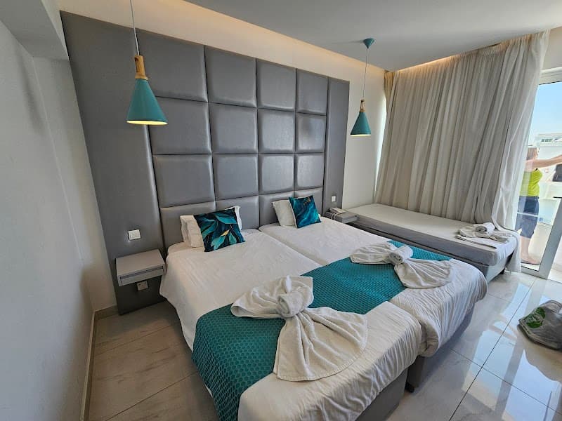 A cozy hotel room at Silver Sands Beach Hotel features modern decor with teal accents, plush bedding, and a balcony view, ideal for travelers seeking comfort and relaxation in Protaras, Cyprus.