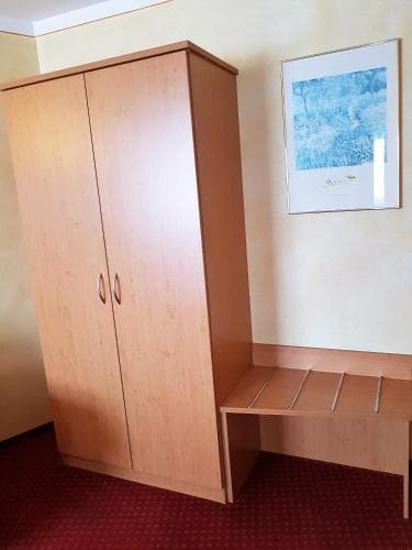 A cozy hotel room corner features a wooden wardrobe and desk set against a warm-toned wall adorned with an abstract painting, ideal for travelers seeking simplicity and functionality.