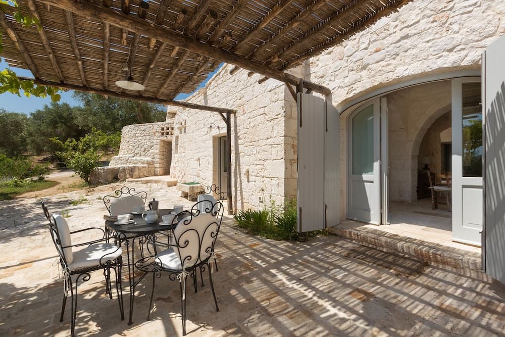 A serene outdoor patio with a rustic stone house, featuring a shaded dining area under a wooden pergola, ideal for travelers seeking a tranquil countryside retreat.