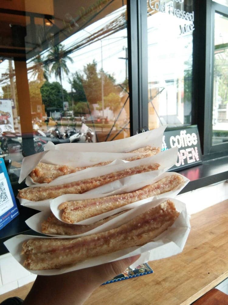 A hand holds freshly fried snacks in front of a cozy café window, reflecting a tropical street scene, perfect for travelers seeking local flavors and a relaxing atmosphere.