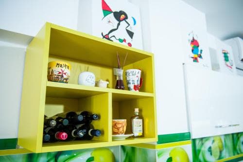 A cozy kitchen corner features a vibrant yellow shelf stocked with wine bottles, olive oil, and decorative mugs, set against playful artwork, ideal for travelers seeking a homely culinary retreat.