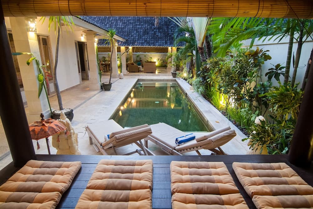 A serene tropical villa courtyard features a tranquil pool surrounded by lush greenery, cozy lounge chairs, and warm lighting, ideal for a relaxing vacation retreat.