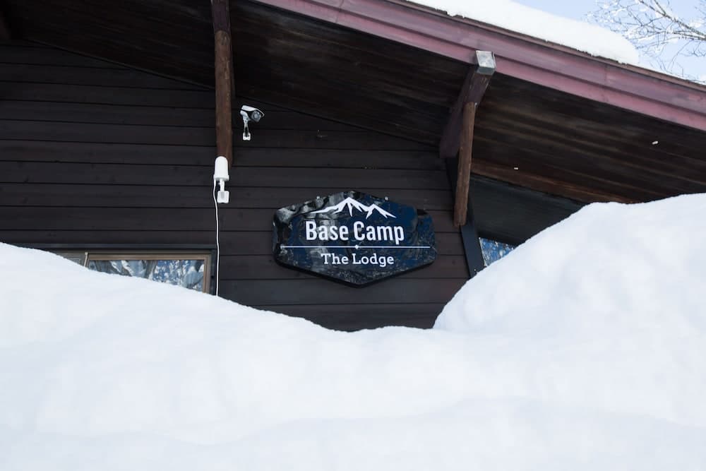 A cozy wooden lodge nestled in deep snow features a "Base Camp The Lodge" sign, ideal for winter retreats and adventure seekers.