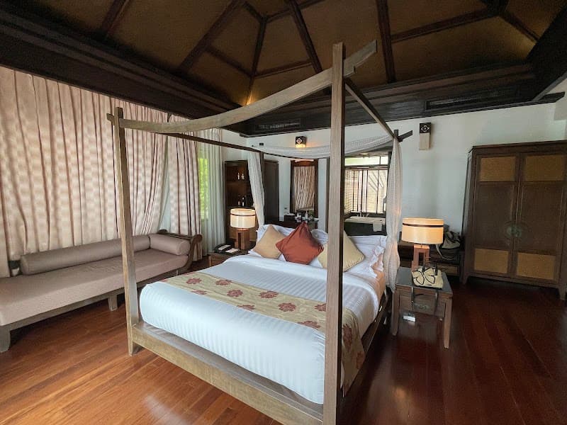 A cozy bedroom at Nora Buri Resort & Spa on Koh Samui exudes tranquility with its wooden four-poster bed, plush furnishings, and natural light, ideal for travelers seeking relaxation and comfort.