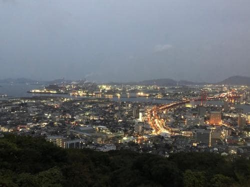 A panoramic evening view of a vibrant coastal city illuminated by twinkling lights, featuring a bridge spanning the waterway, ideal for travelers seeking scenic urban vistas.