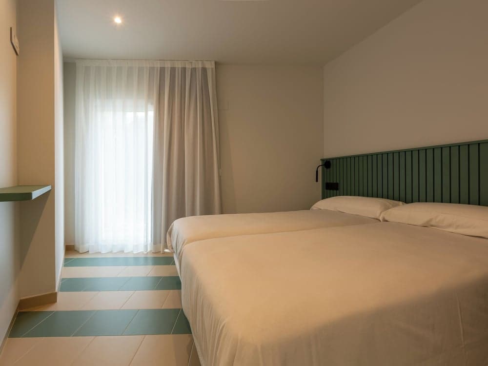 A serene hotel room features two neatly made beds with a green headboard, soft natural light filtering through sheer curtains, and a tiled floor, ideal for travelers seeking comfort and simplicity.