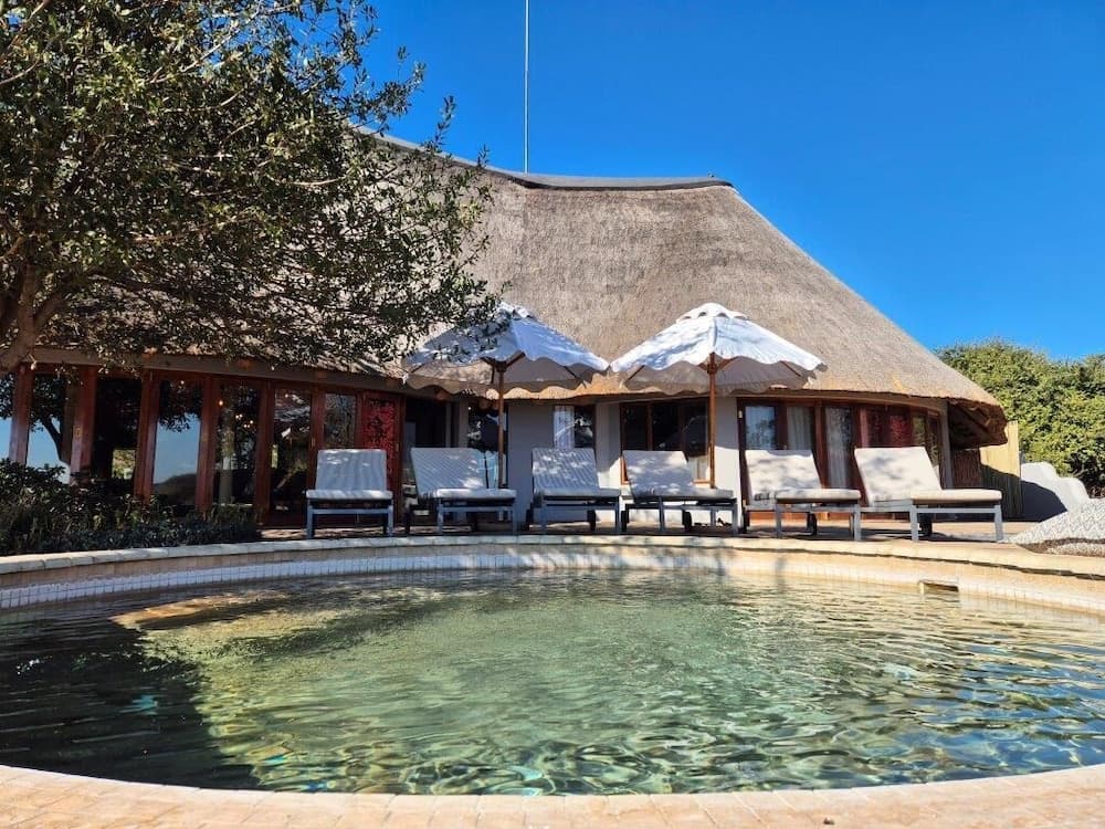 A serene tropical lodge features a tranquil poolside area with lounge chairs under white umbrellas, set against a thatched-roof building and lush greenery, ideal for a relaxing vacation getaway.