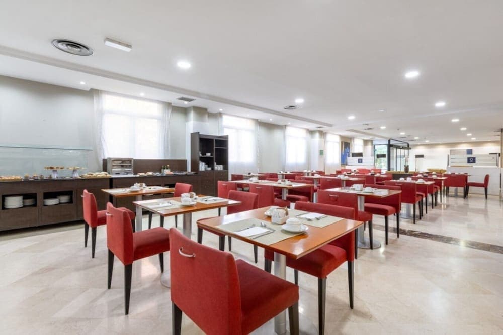 A spacious hotel dining area with red chairs, wooden tables, and a buffet setup bathed in natural light from large windows, ideal for travelers seeking a comfortable breakfast or lunch spot.